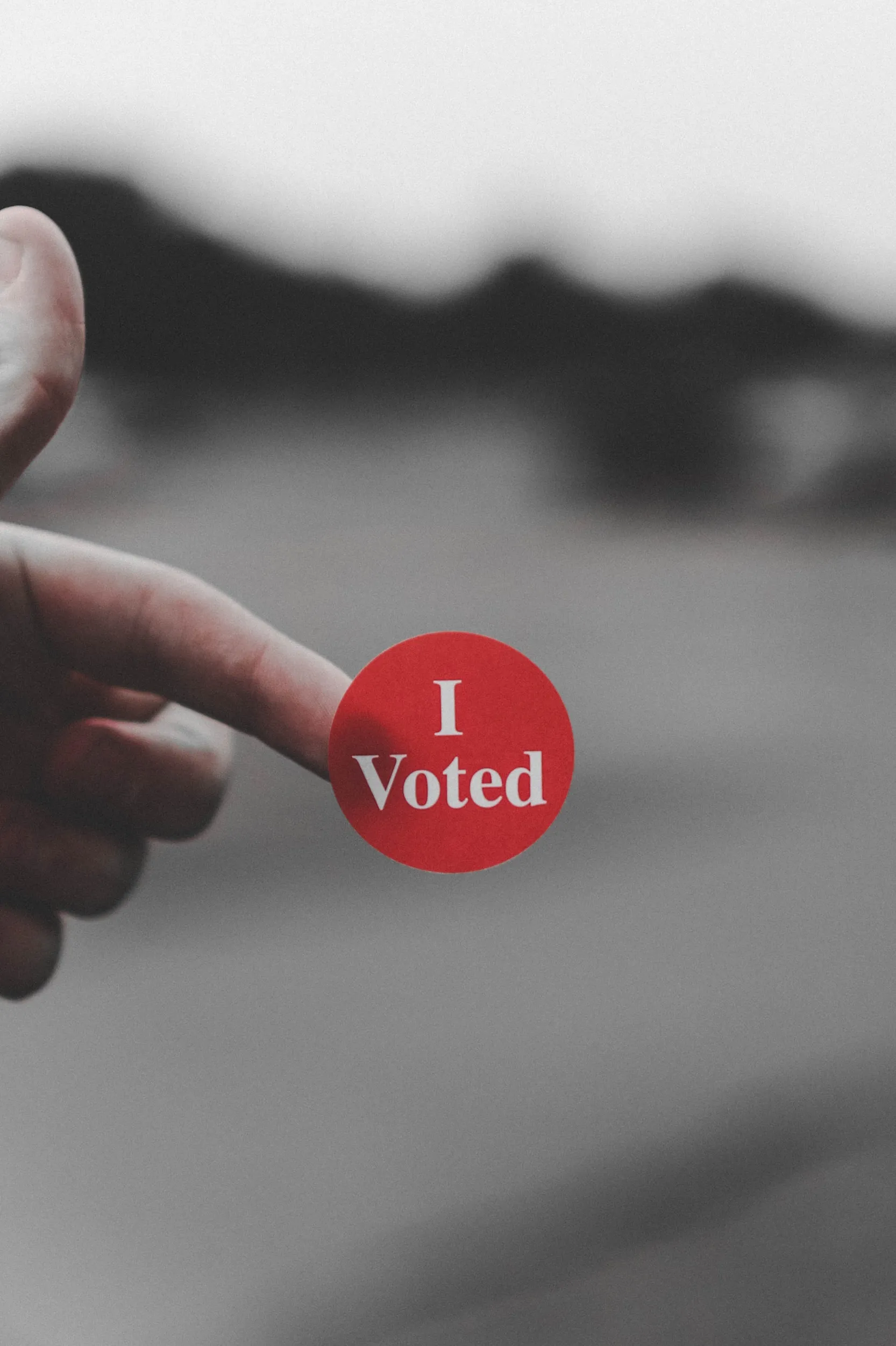 A finger holding an I Voted sticker, with selective red color against a muted background